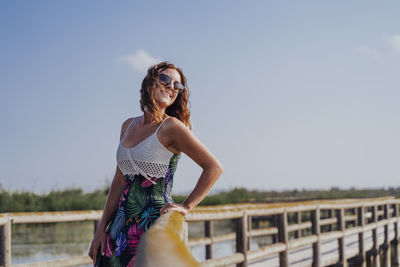 Smiling woman standing by railing on footbridge against sky