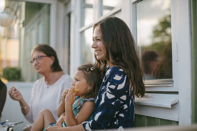 Smiling mother with daughter eating fruit while sitting by senior woman at porch