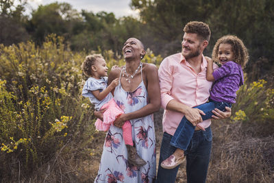 Happy parents carrying cute daughters while standing on field against trees in park