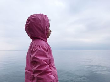 Side view of girl wearing hooded shirt while standing against sea