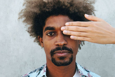 Anonymous friend covering eye of crop stylish ethnic male with afro hairstyle looking at camera near concrete wall