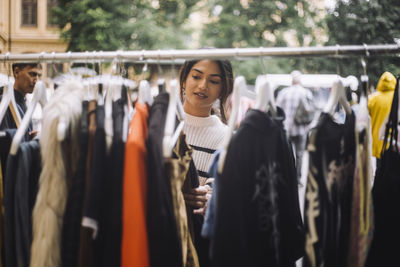 Young woman selecting dress hanging on clothes rack at flea market