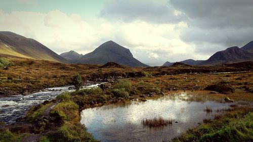 Scenic view of stream on landscape by mountains against cloudy sky