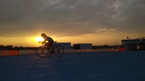 Silhouette man riding bicycle on street against sky during sunset