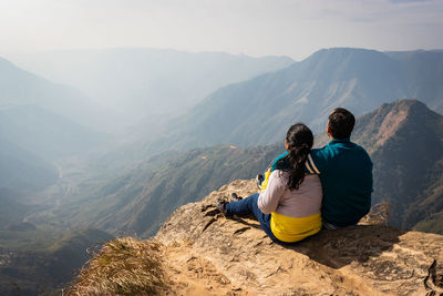 Rear view of couple sitting on cliff against mountains
