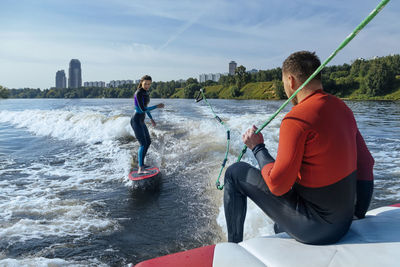 Man sitting at edge of moving boat looking at female wakeboarder surfing behind