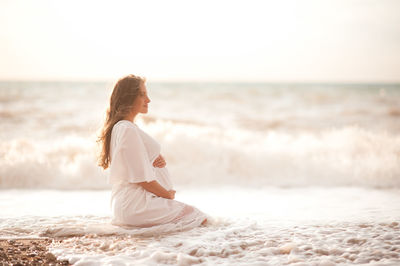 Beautiful pregnant woman wear white elegant comfy dress hold tummy sitting in sea shore over nature