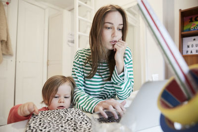 Curious girl touching book while working mother using laptop at table