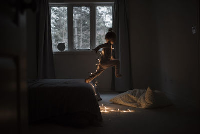 Side view of girl with illuminated string lights jumping from bed at home