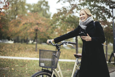 Smiling businesswoman enjoying music while walking with bicycle during autumn