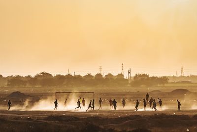 People playing soccer on field against sky during sunset