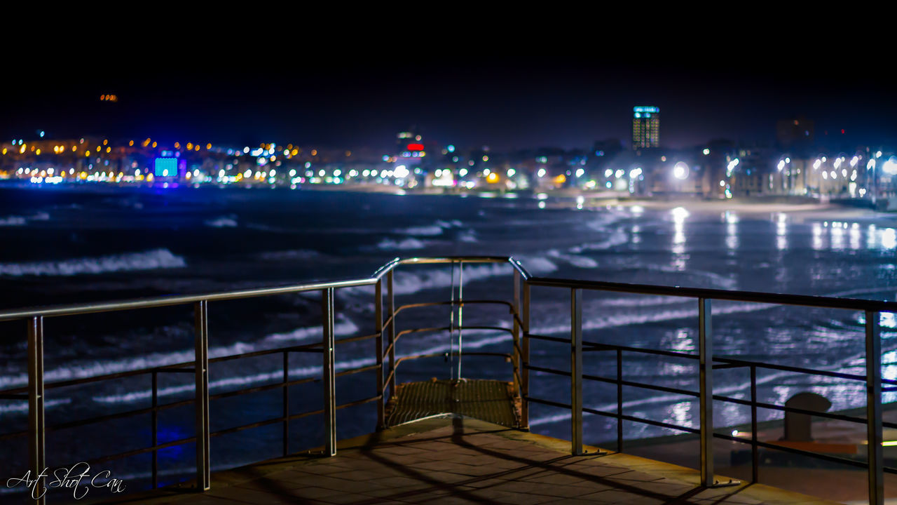 ILLUMINATED BUILDINGS BY SEA AGAINST SKY AT NIGHT