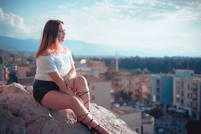 Full length of beautiful young woman sitting on rock in city against sky