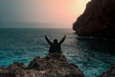 Rear view of man sitting on rock by sea against sky