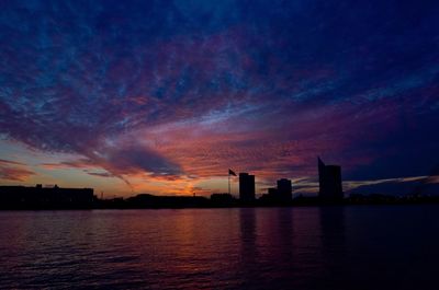 Silhouette buildings by sea against sky during sunset