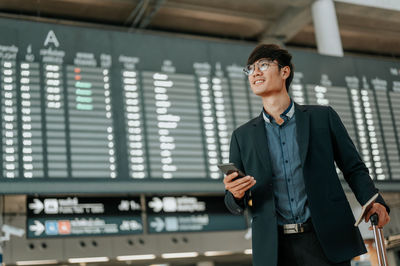 Businessman using mobile phone while holding suitcase and passport at airport