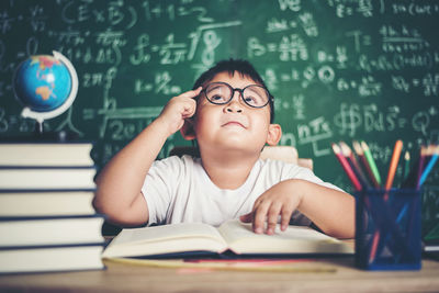 Cute boy studying at table against blackboard