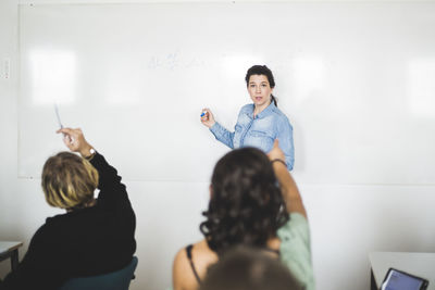 Students pointing at teacher standing against whiteboard in classroom