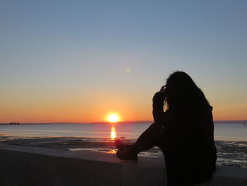 Woman sitting on retaining wall by beach during sunset