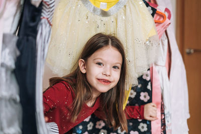 Smiling tween little girl with long dark hair in red dress among dresses in wardrobe at home