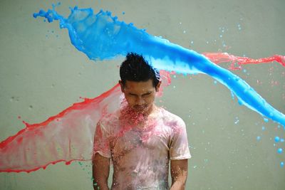 Color water splashing over young man standing against gray background