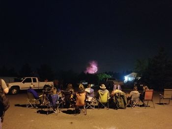 People sitting on illuminated street against sky at night