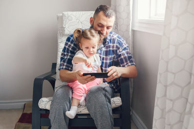 Man with daughter using phone on chair at home