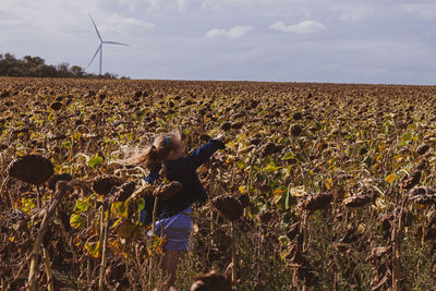 Blond hair child girl in denim jacket walks in sunflowers field wind turbines background. eco energy