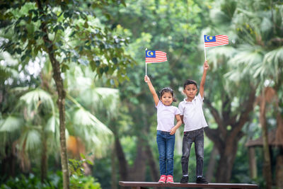 Portrait of siblings holding malaysian flag while standing on bench against trees at park