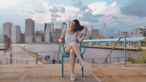 Portrait of young woman sitting on chair at harbor with cityscape in background against sky