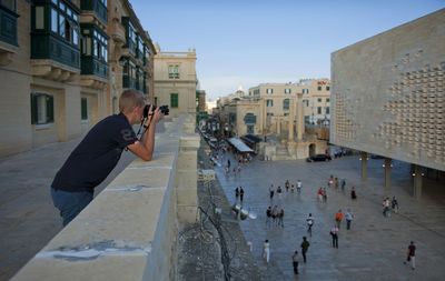 Side view of man photographing people on footpath amidst buildings in city