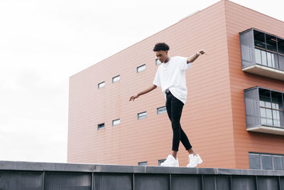 From below side view of young stylish african american male looking away in white shirt and tight pants with white sneakers standing on fence against pink city building