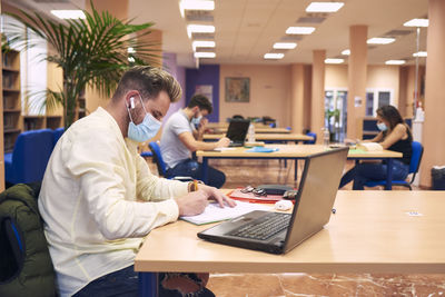 A young boy with a mask sitting and working with his laptop in the library