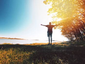 Rear view of man with arms outstretched standing on grassy field by river during sunny day