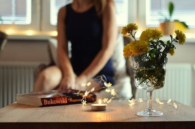 Flower vase by lit tea light candle on table while woman in background