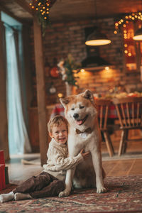 Candid authentic happy little boy in knitted beige sweater hugs dog with bow tie at home on xmas