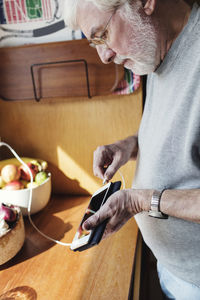 Senior man connecting cable to mobile phone at kitchen counter on sunny day