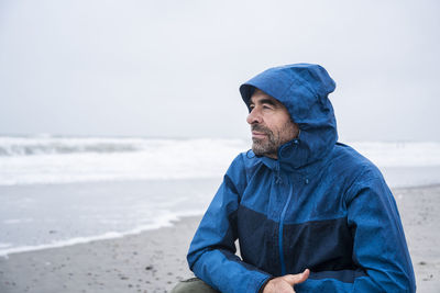 Contemplated mature man in blue raincoat crouching at beach against sky