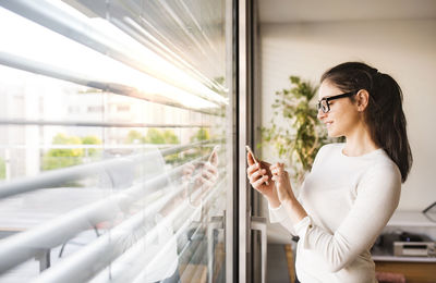 Smiling woman standing at window at home using smartphone