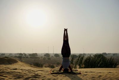 Rear view of woman practicing headstand against sky during sunset