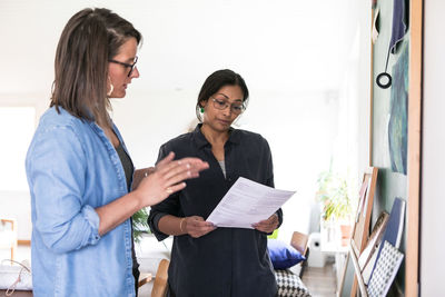 Female design professionals discussing paper while standing in home office