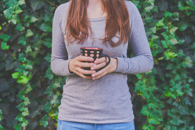 Midsection of woman holding coffee cup while standing against plants