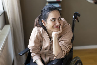 Smiling young woman with hand on chin sitting on wheelchair at home