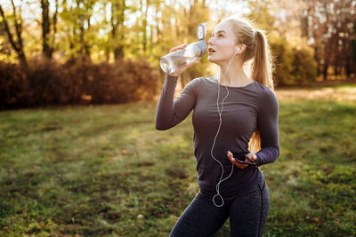 Fitness in the park, girl drinking water, holding smartphone and headphones in her hand.