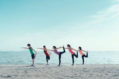 Young friends practicing yoga in warrior position on shore at beach during sunny day
