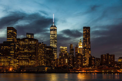 Illuminated one world trade center amidst buildings in front of sea at dusk