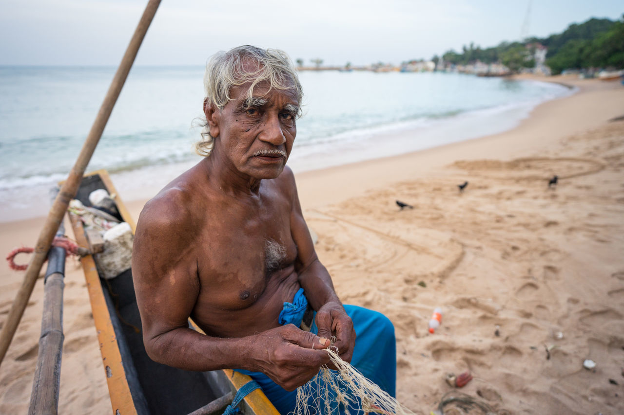 Tangalle, sri lanka - july 28, 2019: side view of ethnic old fishermen in casual light wear unwinding fishing net on big yellow boat at tropical sandy beach
