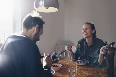 Happy woman with mobile phone looking at man while eating pasta at home