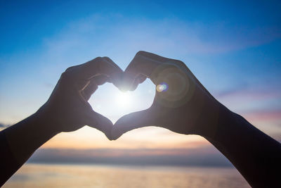 Close-up of hand making heart shape against sky during sunset