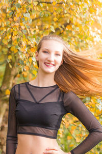 Portrait of young beautiful woman tossing her long blond hair while standing by autumn tree